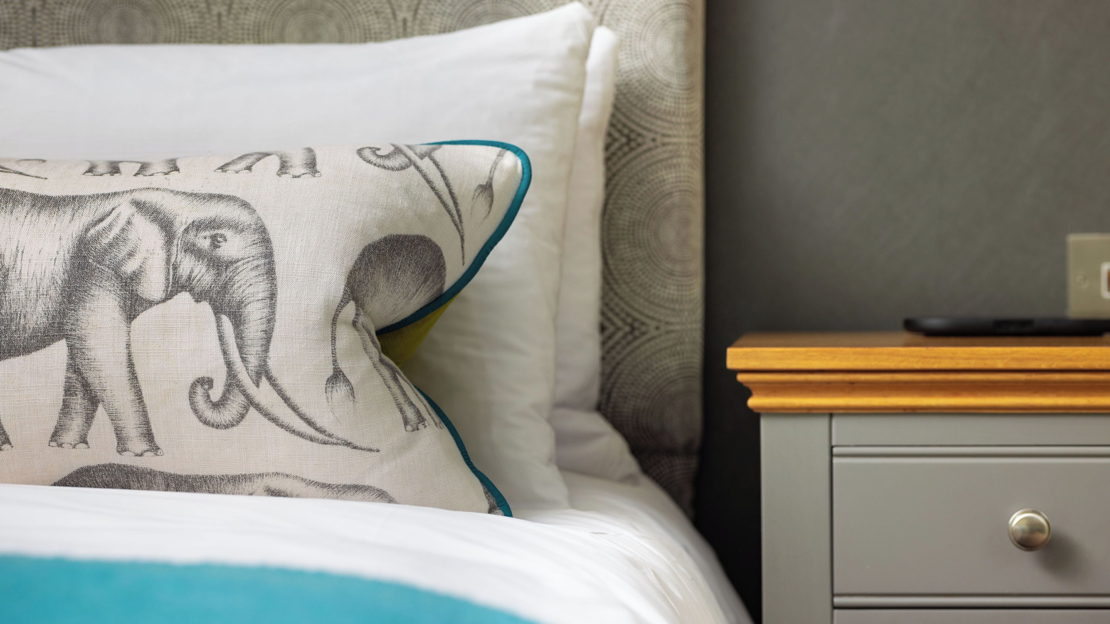 A bed with a white pillow featuring an elephant design, a wooden nightstand with a remote control, and a gray drawer.