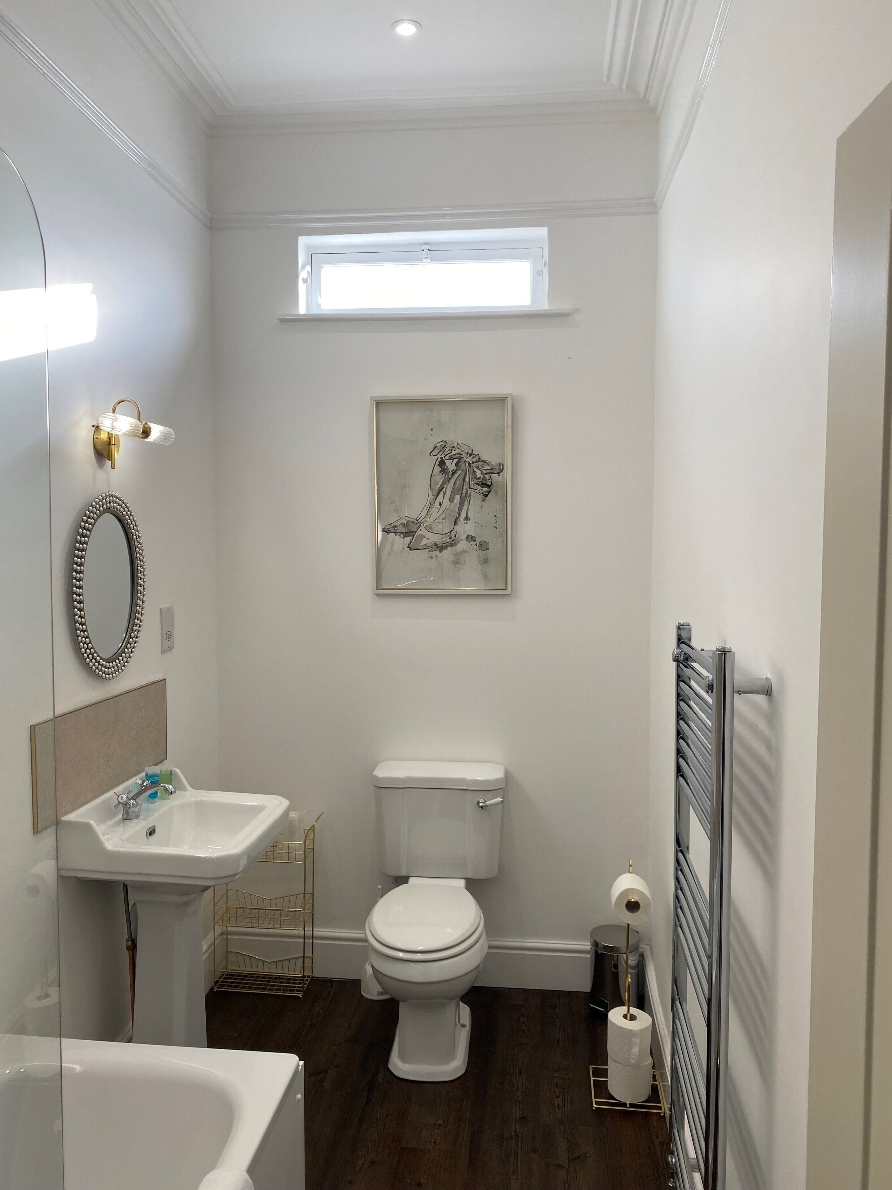 Une salle de bain moderne avec un lavabo, des toilettes et une baignoire, tous de couleur blanche. Il y a une fenêtre au-dessus des toilettes et une œuvre d’art encadrée au mur.