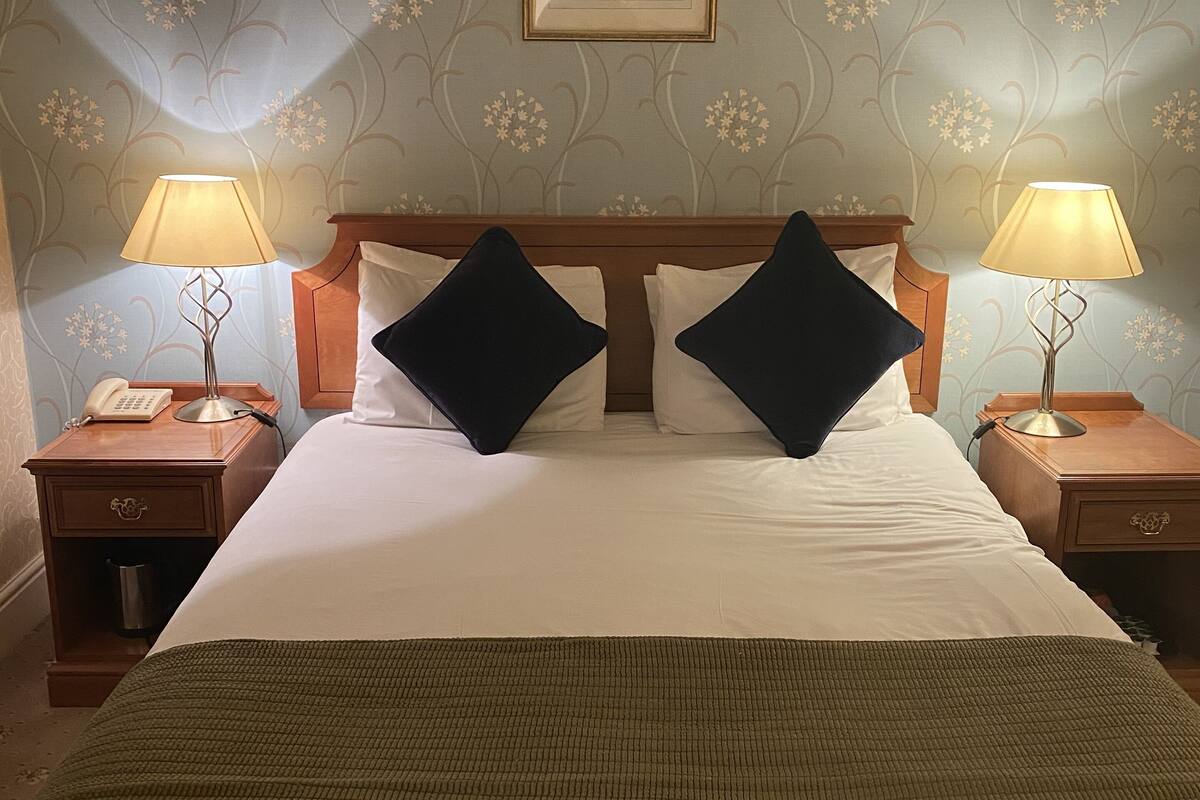 A neatly made bed with two black pillows, flanked by nightstands with lamps, in a room with floral wallpaper and a framed picture above the bed.