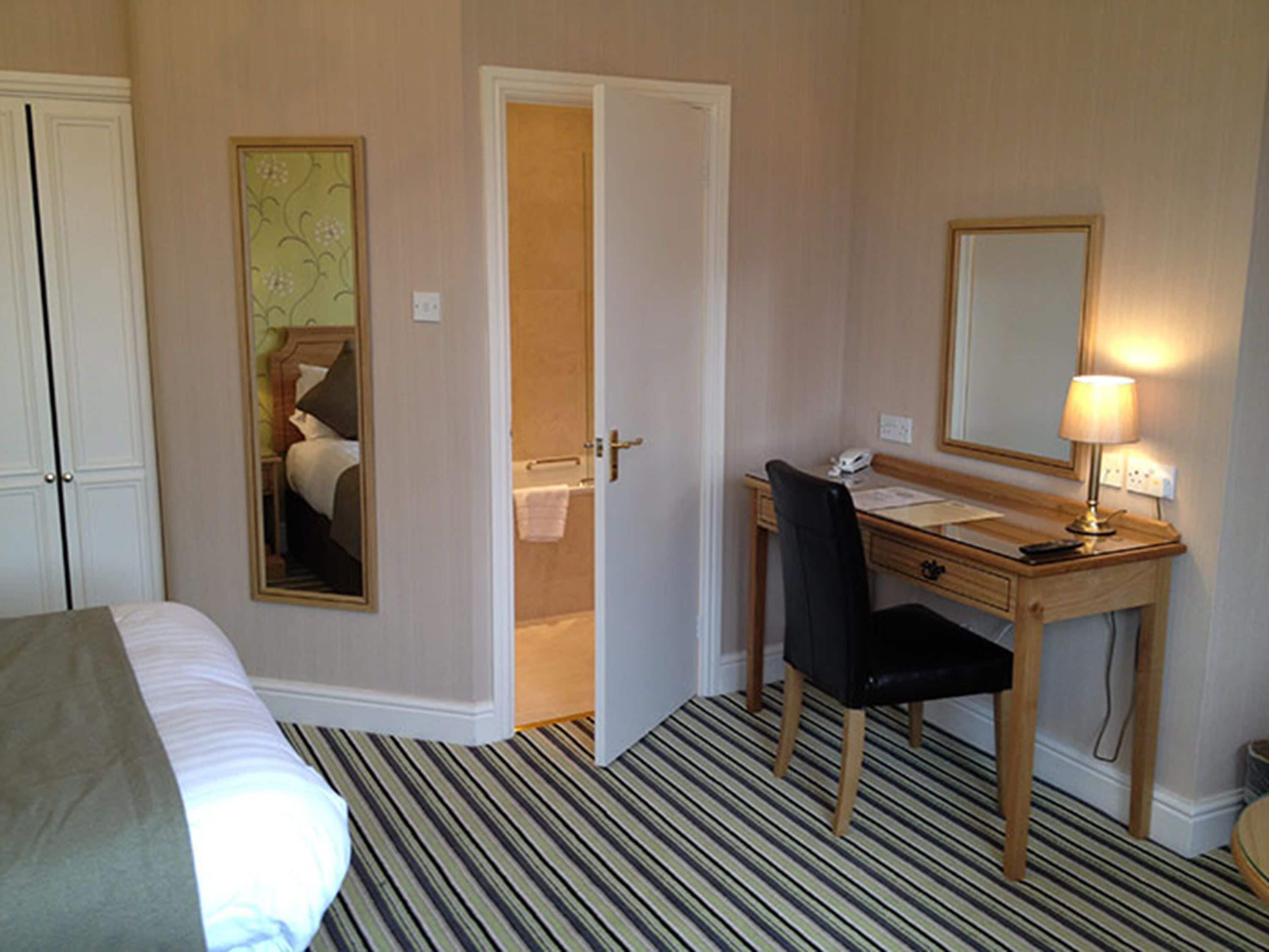 A hotel room with a bed, a desk with a chair, a mirror, and a lamp.