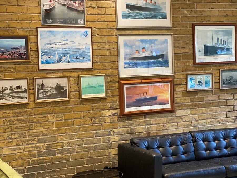 A room with a brick wall adorned with framed maritime paintings, a black leather sofa, and a small round table.