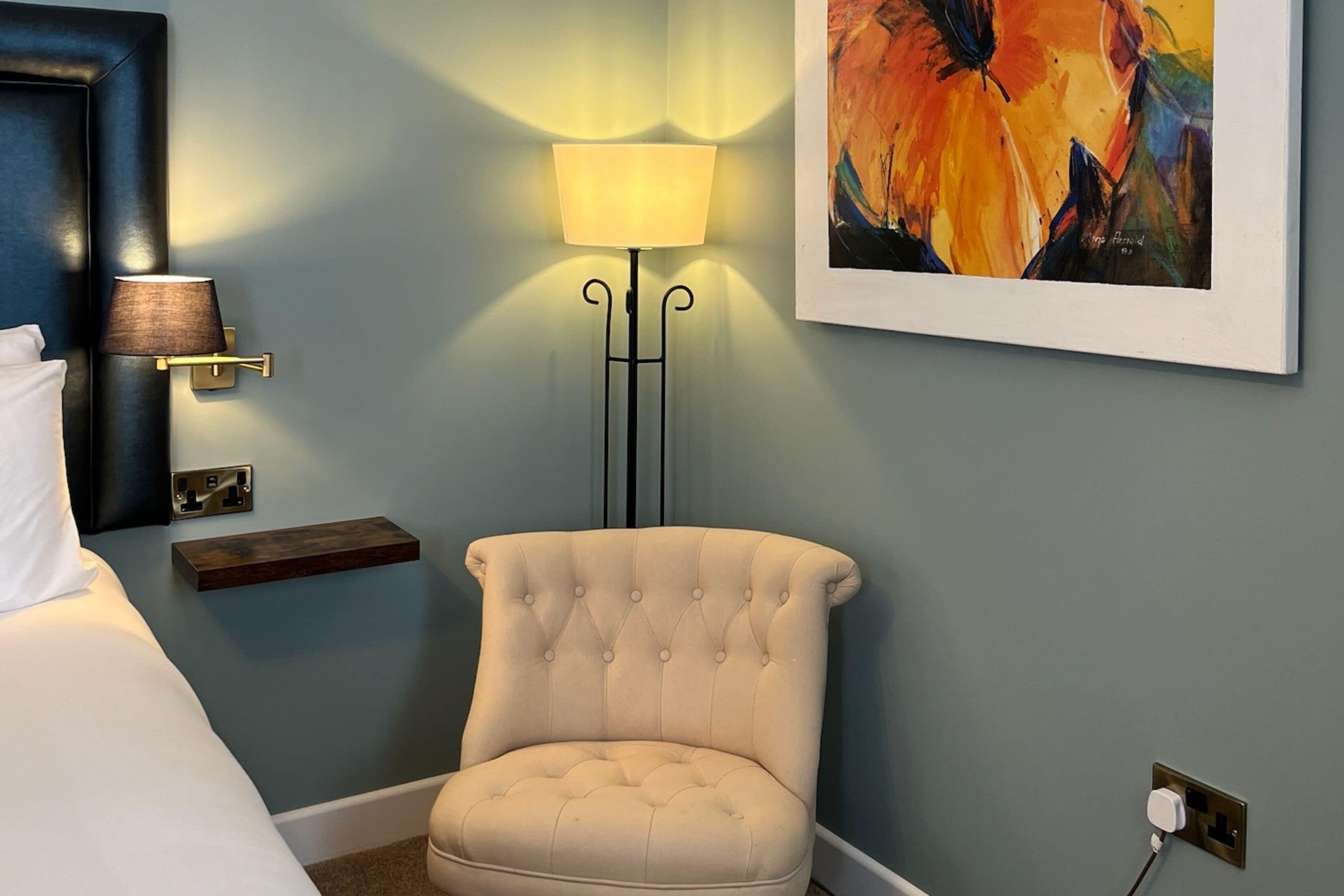 Un dormitorio con un sillón capitonado, una mesita de noche con lámpara, un cuadro en la pared y un enchufe.
