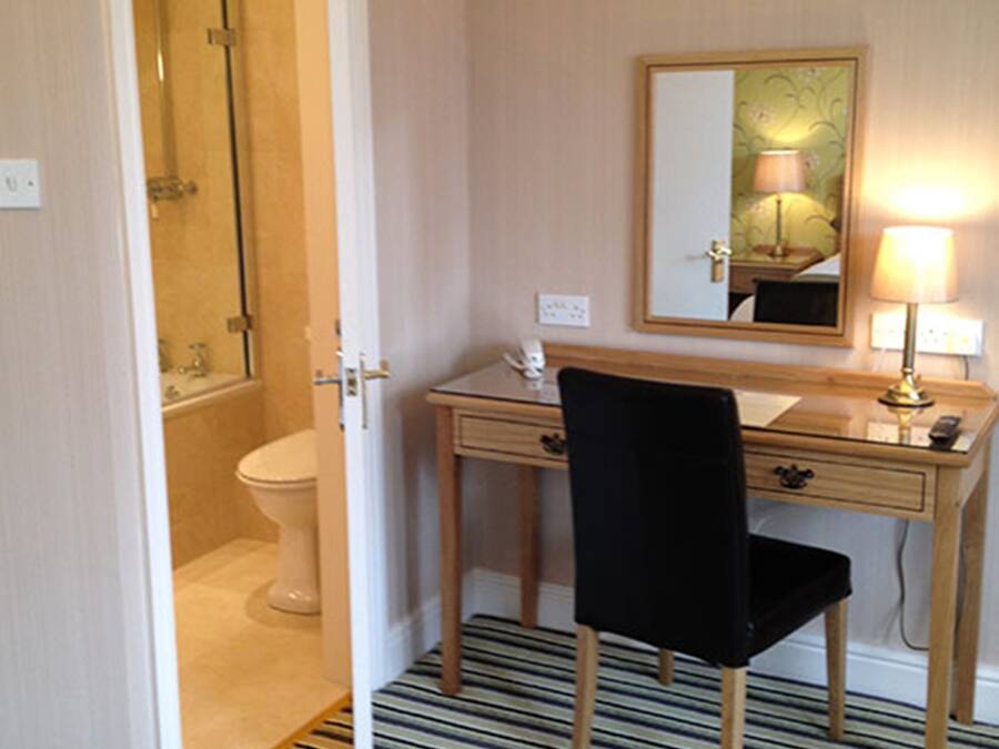 A hotel room with a bathroom, a desk with a chair, a mirror, and a lamp.