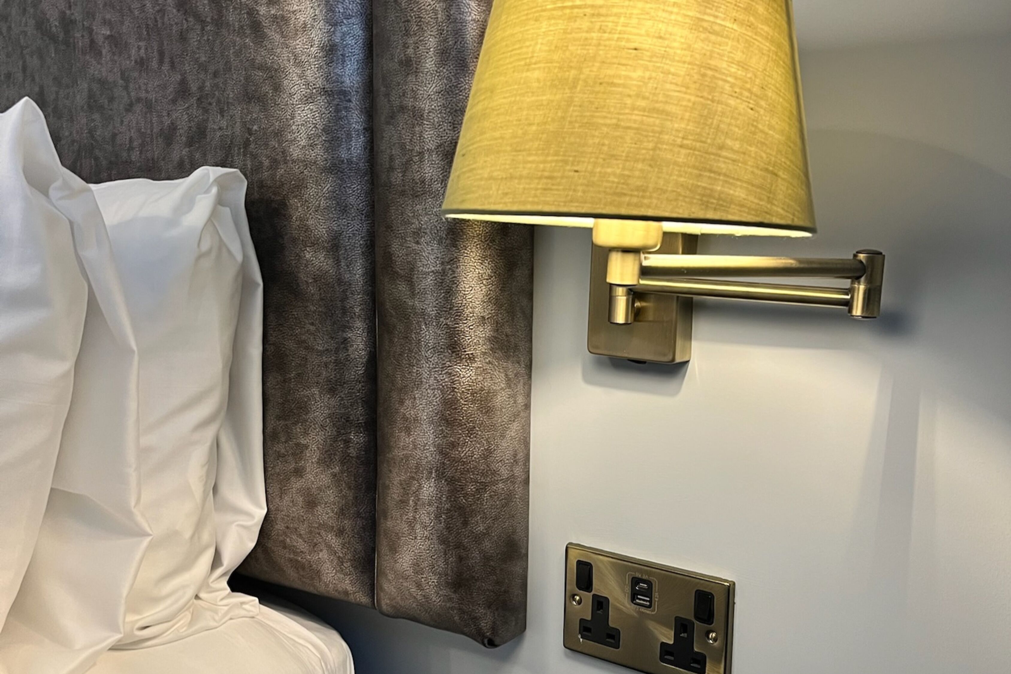 Une lampe murale avec un abat-jour en tissu, une prise de courant et un oreiller blanc sur un lit.
