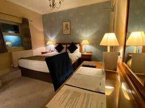 A hotel room with a bed, two bedside tables with lamps, a chair, a desk with papers, and a window with curtains.