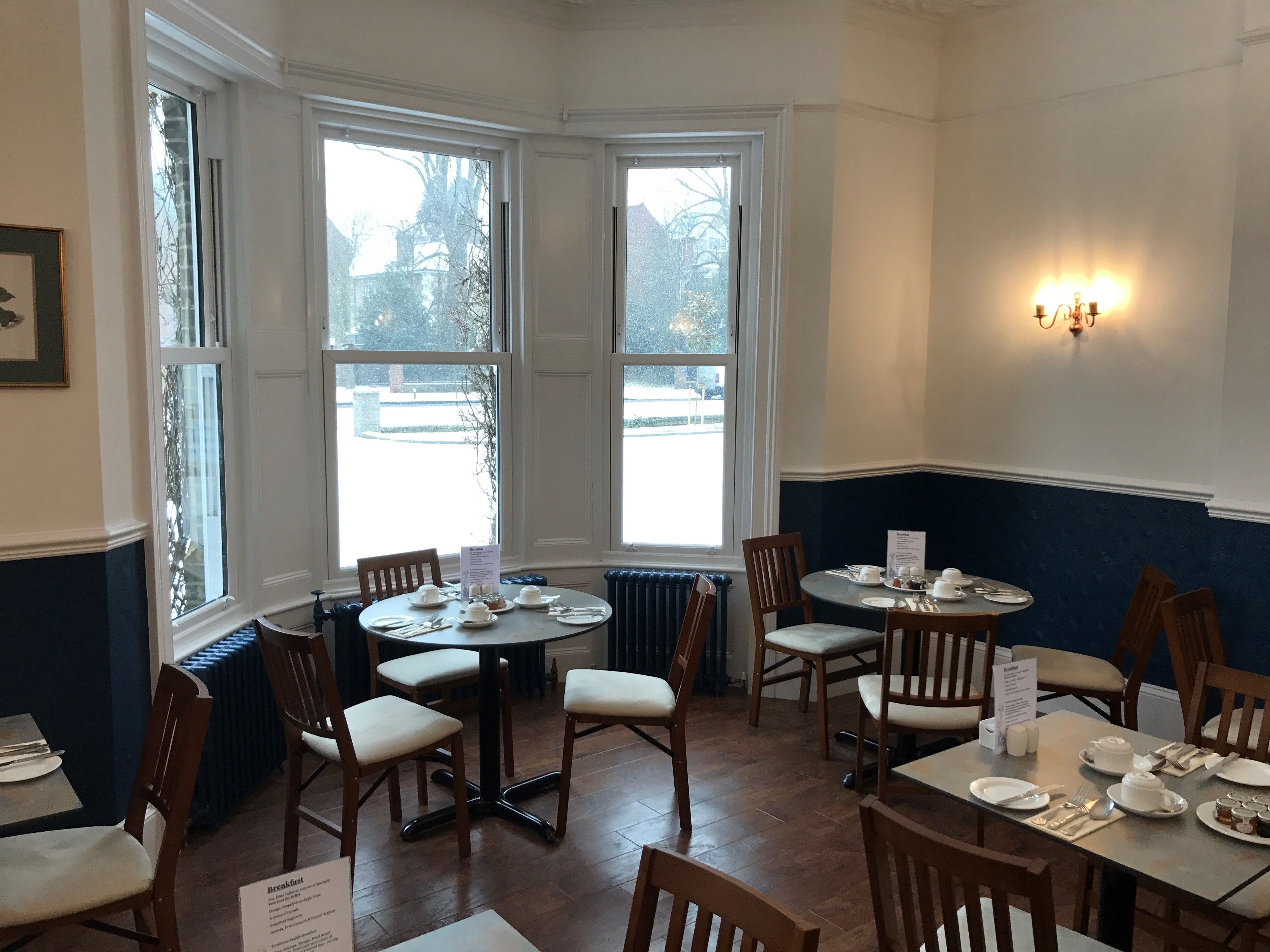 A dining area with round tables and chairs, large windows with a view of the outside, and a wall-mounted light fixture.