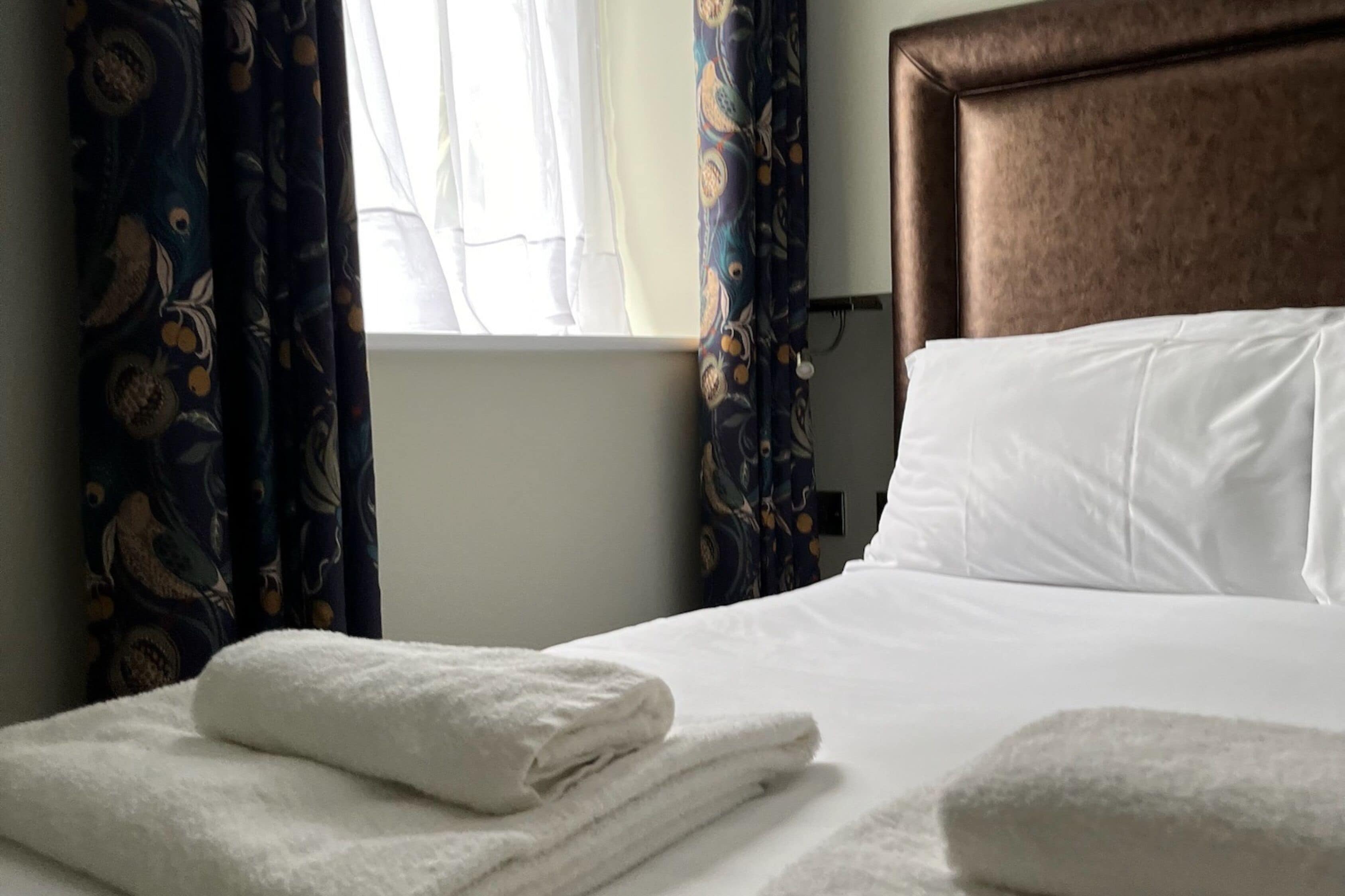 A neatly made bed with white linens and folded towels, a window with sheer curtains, and a brown headboard.