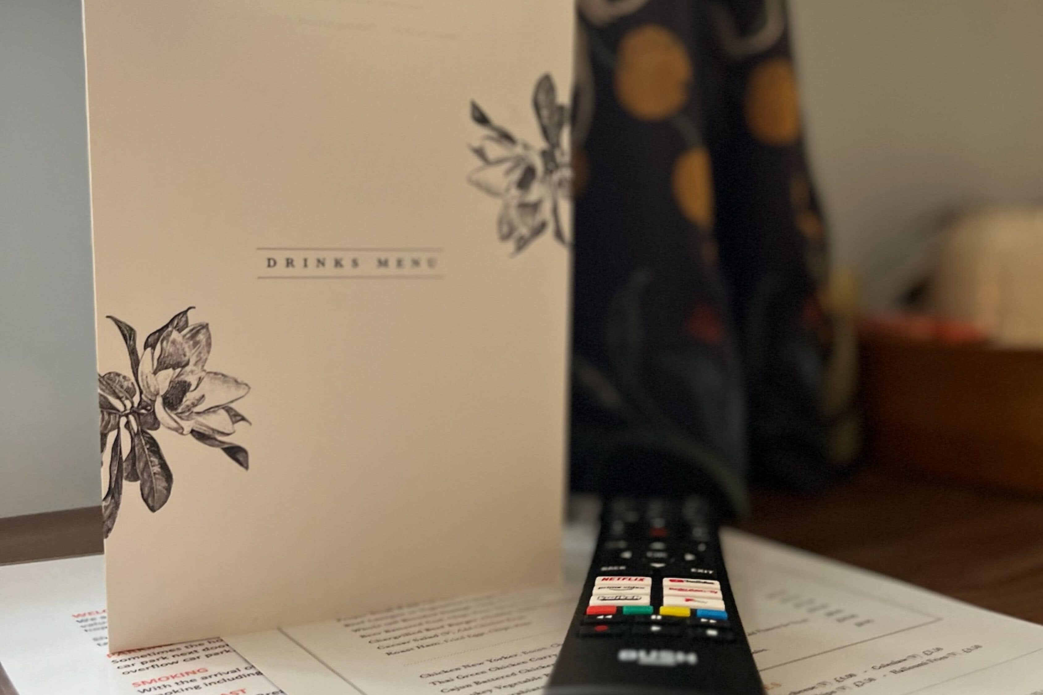 A remote control with various buttons, a menu card with a floral design, and a blurred background with a curtain.
