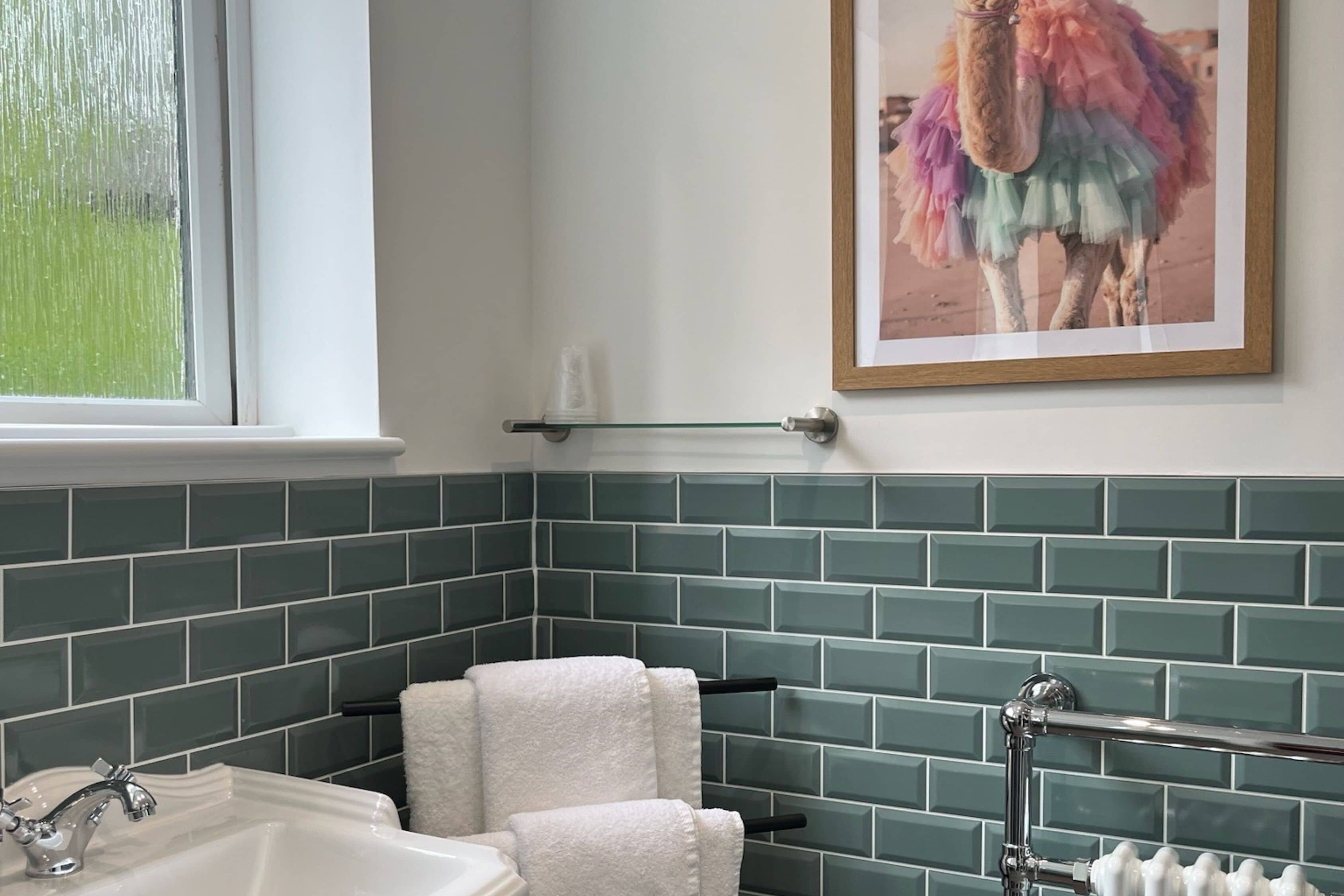 A bathroom with green tiled walls, a white sink, and a framed picture of a camel.