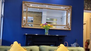 Een kamer met een blauwe wand, een open haard, twee groene fauteuils met gele kussens, en een grote spiegel boven de open haard.