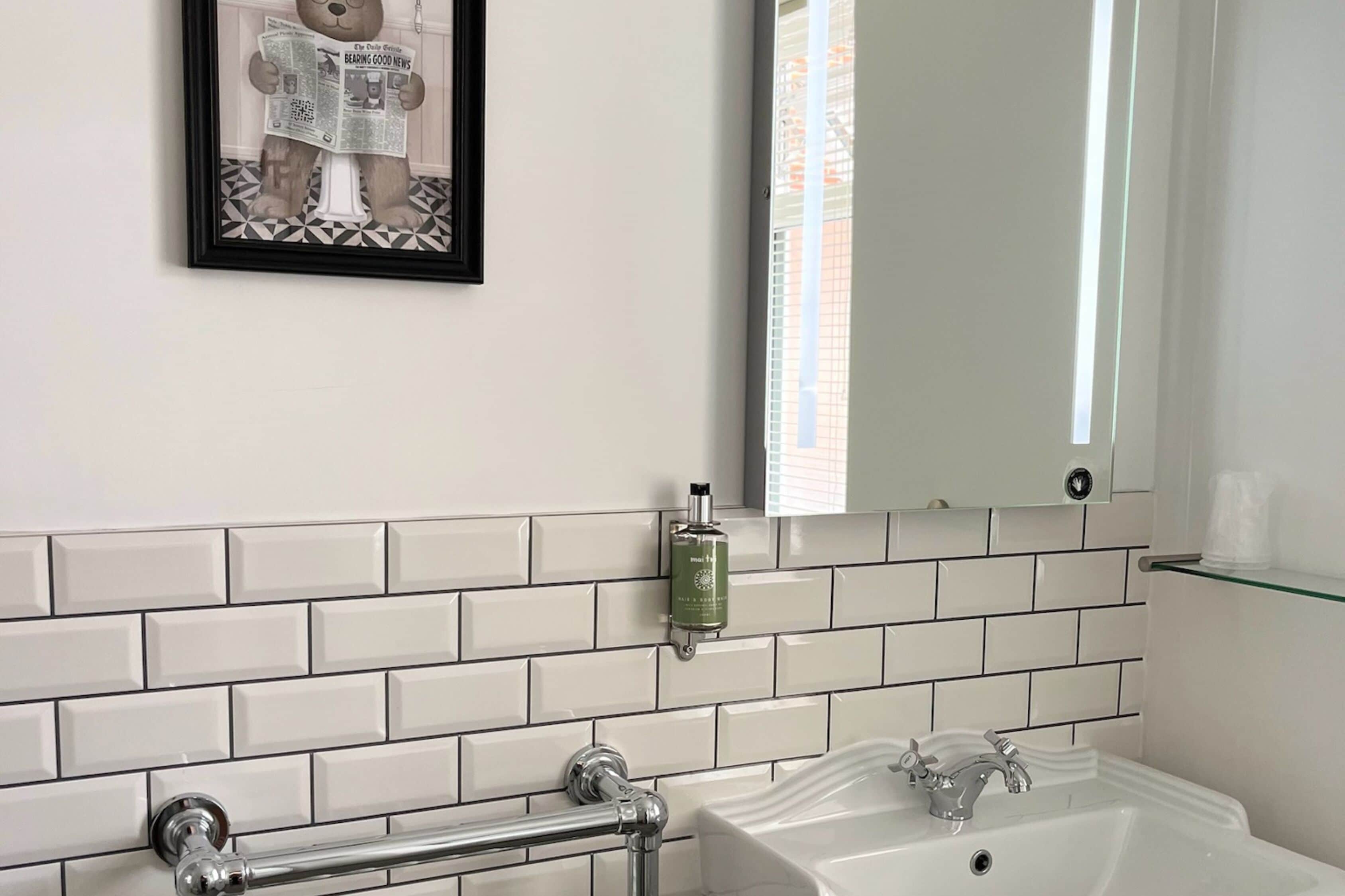 Un baño con baldosas blancas tipo metro, un cuadro enmarcado de un oso leyendo un periódico, una botella verde de jabón de manos y un estante de vidrio con tazas.