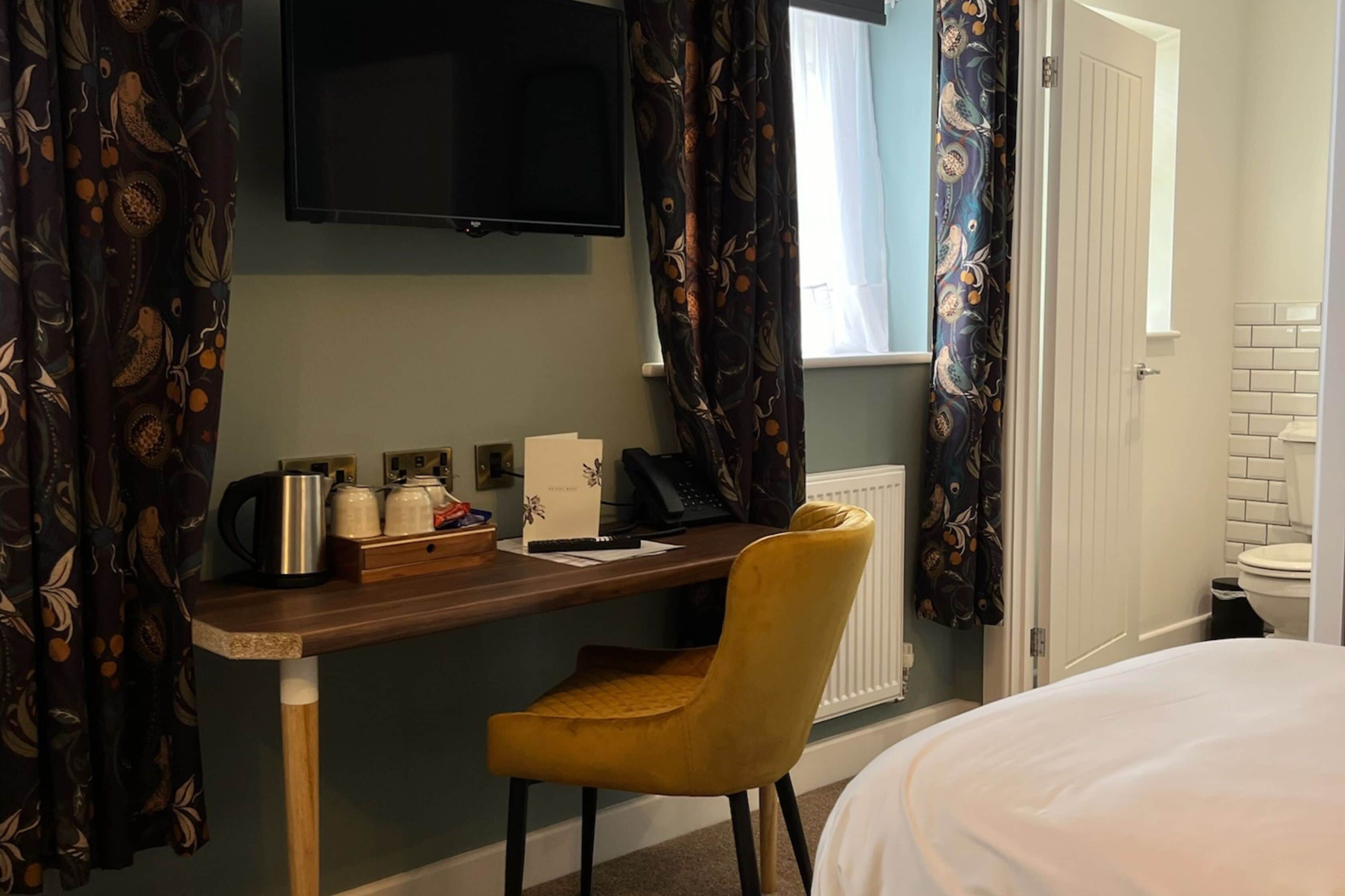 A hotel room with a wooden desk, a yellow chair, a bed, a television, and a window with curtains.