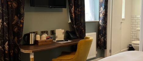 A hotel room with a wooden desk, a yellow chair, a bed, a television, and a window with curtains.