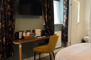 A hotel room with a wooden desk, a yellow chair, a bed, a television, and a window with curtains.