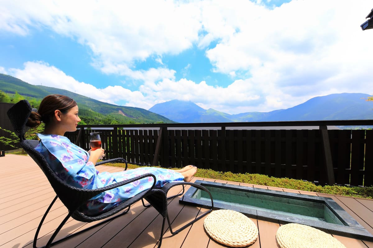 A person relaxing on a deck chair with a view of mountains and a pool.