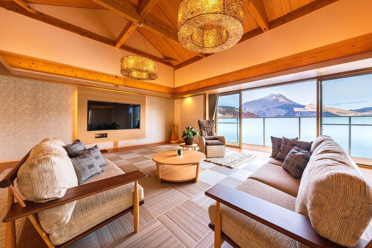 A spacious living room with a large TV, comfortable seating, a coffee table, and a view of a mountain and lake.