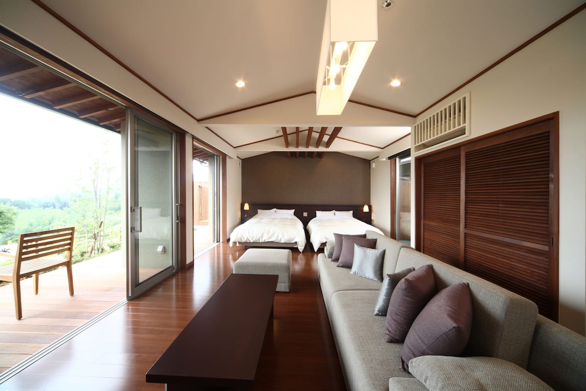 A modern hotel room with a large bed, a sofa, a bench, and a view of the outdoors.