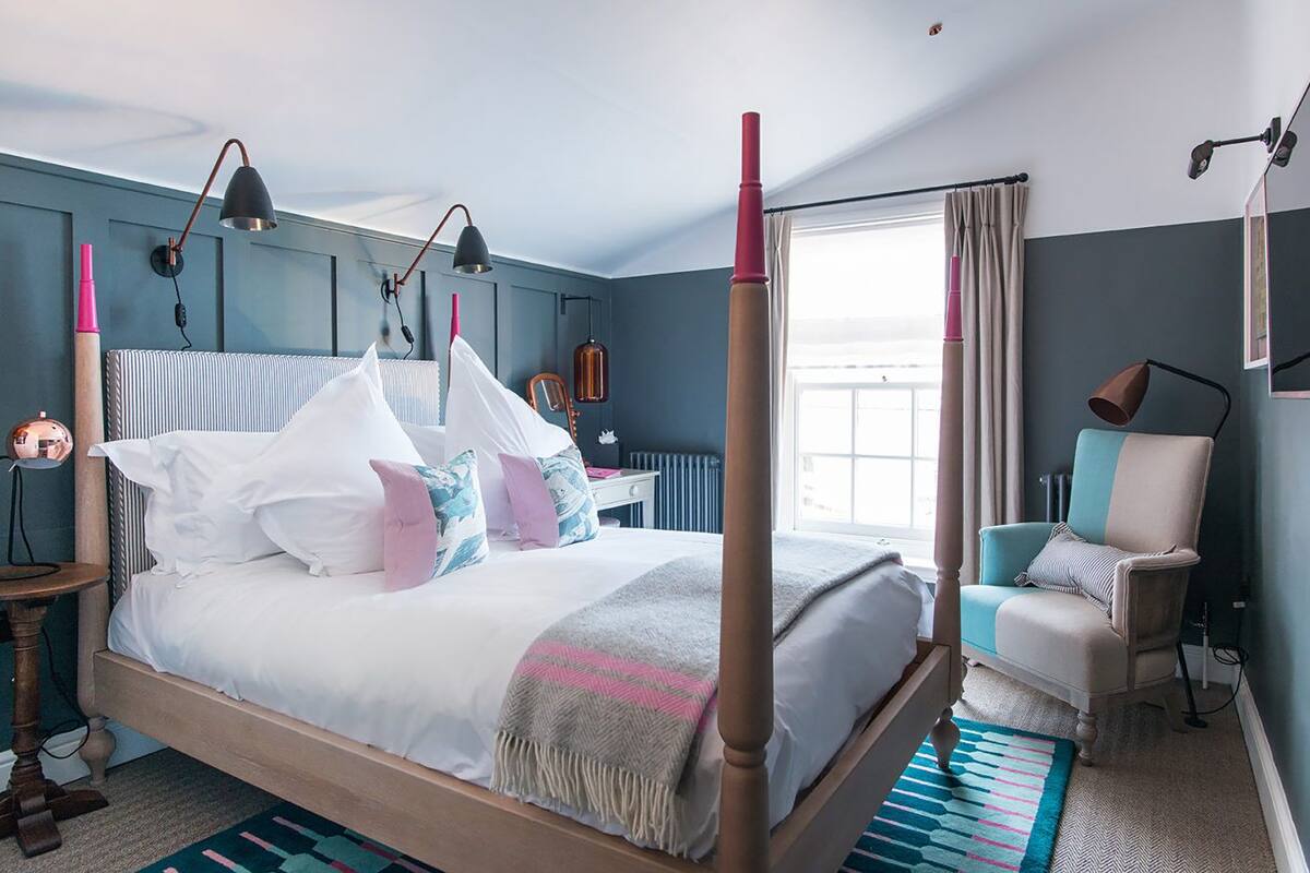 A four-poster bed with white linens and multiple pillows, a bedside table with a lamp, a chair with a striped cushion, and a window with curtains.