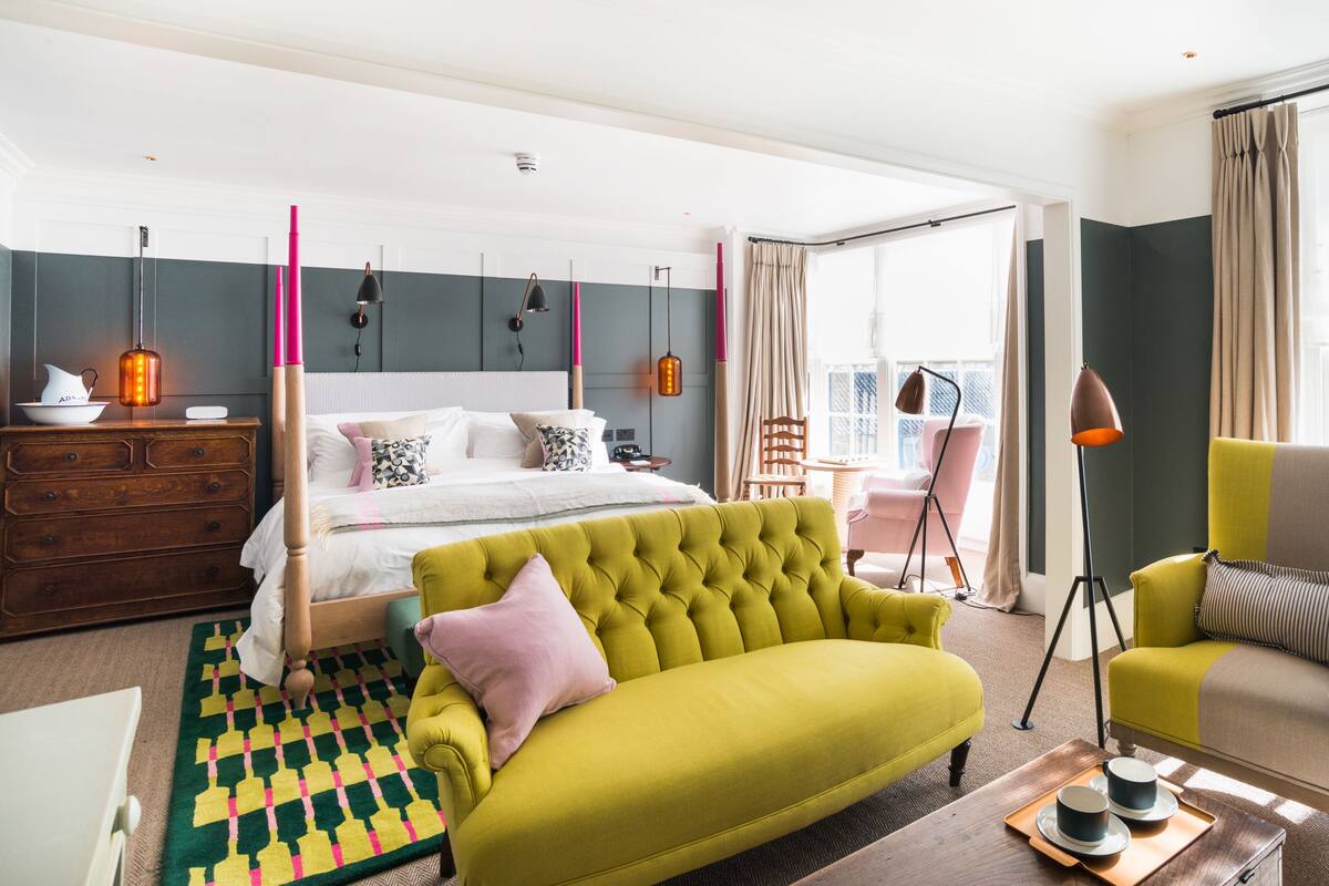 A modern hotel room with a large bed, a green sofa, a chair, and a dining area with a tray of cups.
