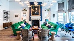 Un vaste espace salon avec des canapés verts, des fauteuils en bois et une table centrale. Un grand tableau est accroché au-dessus d’une cheminée, et la pièce est dotée de grandes fenêtres ornées de rideaux à rayures.