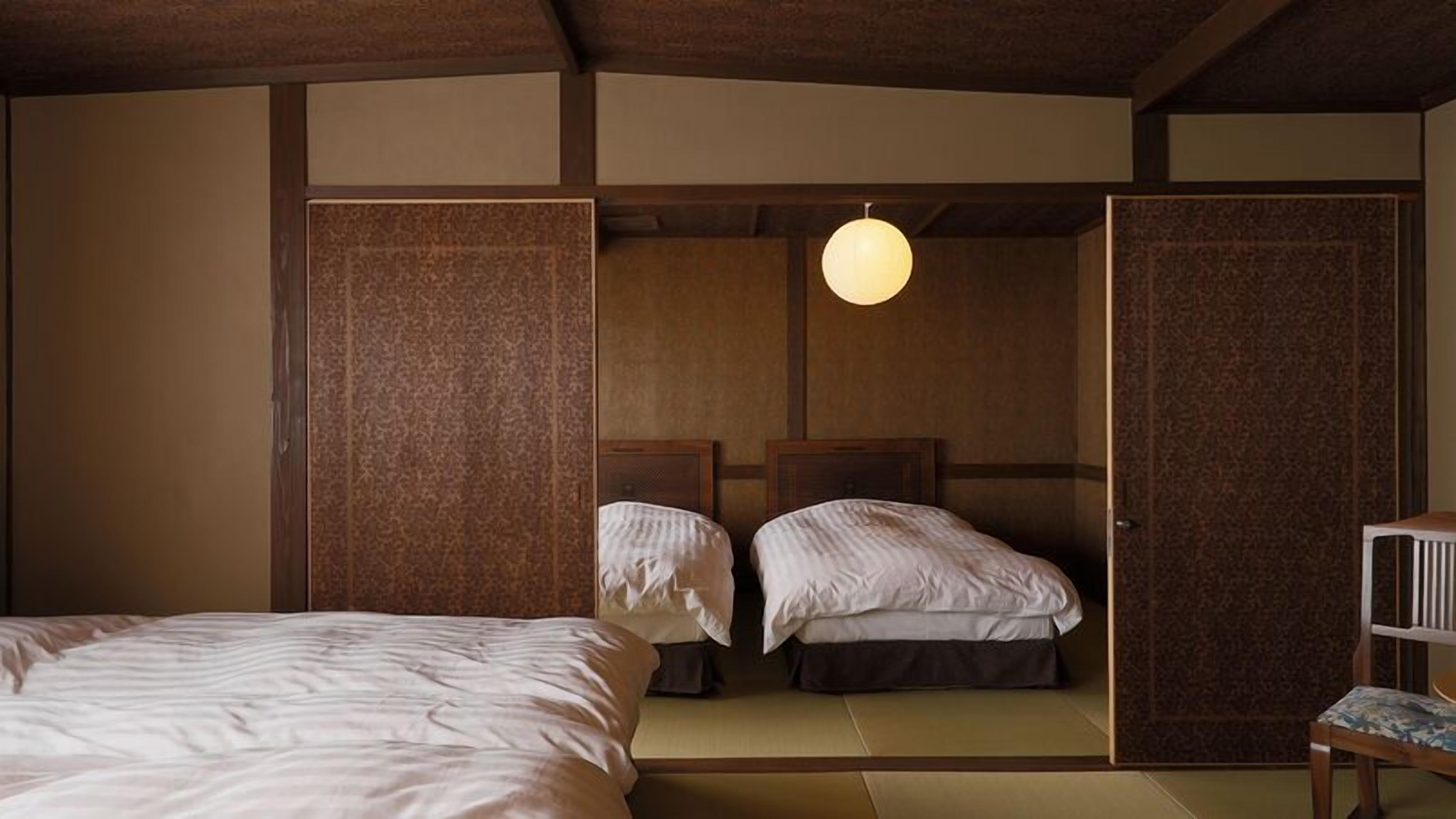 A traditional Japanese-style room with tatami flooring, two beds with white bedding, a hanging light fixture, and wooden sliding doors.