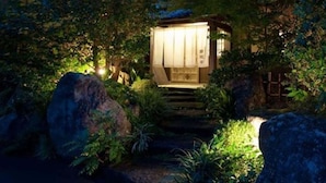 Ein traditionelles japanisches Holzgebäude mit kleinem Eingang, umgeben von üppigem Grün und großen Steinen.