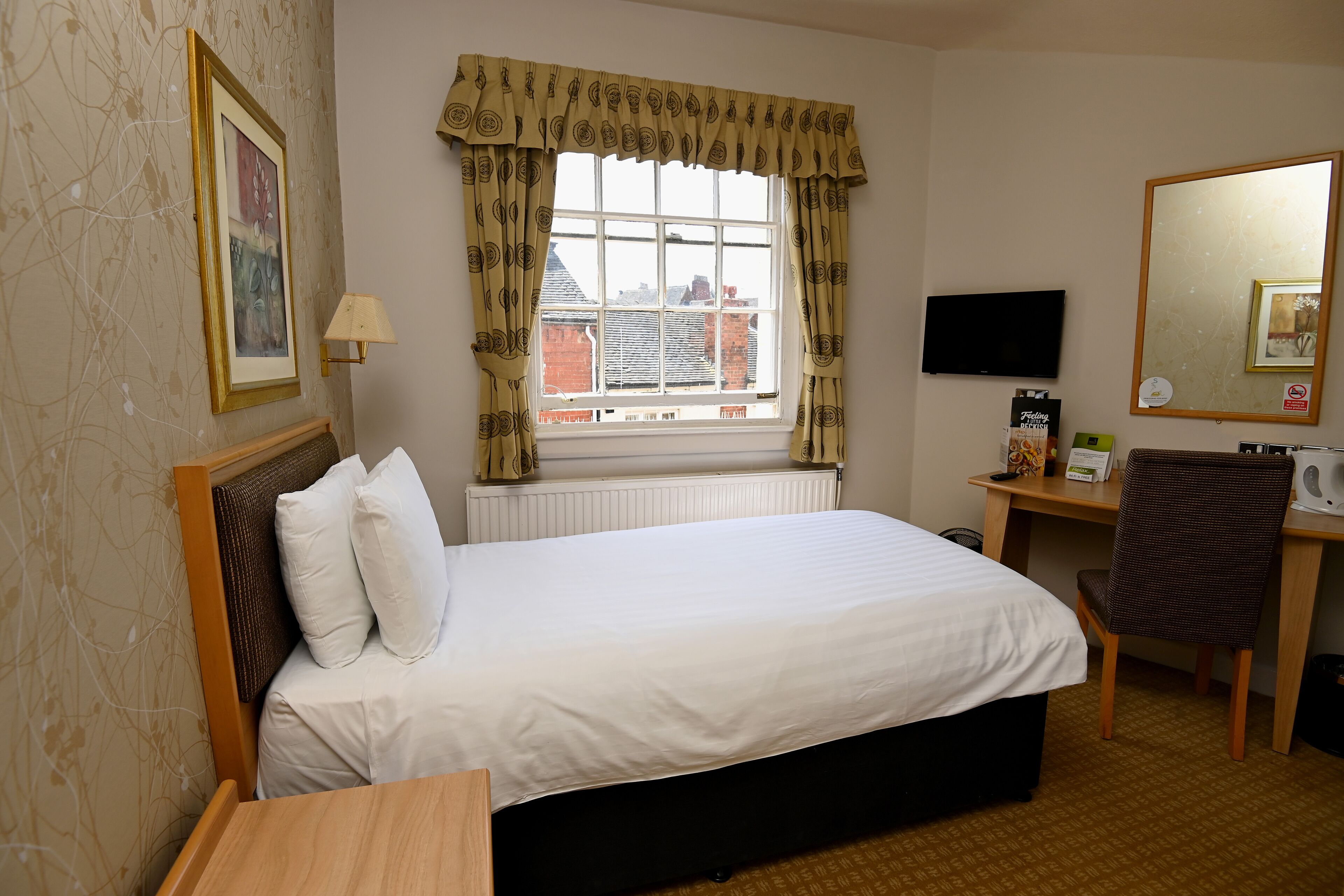 A hotel room with a bed, a desk, a chair, a TV, a mirror, and a window with curtains.