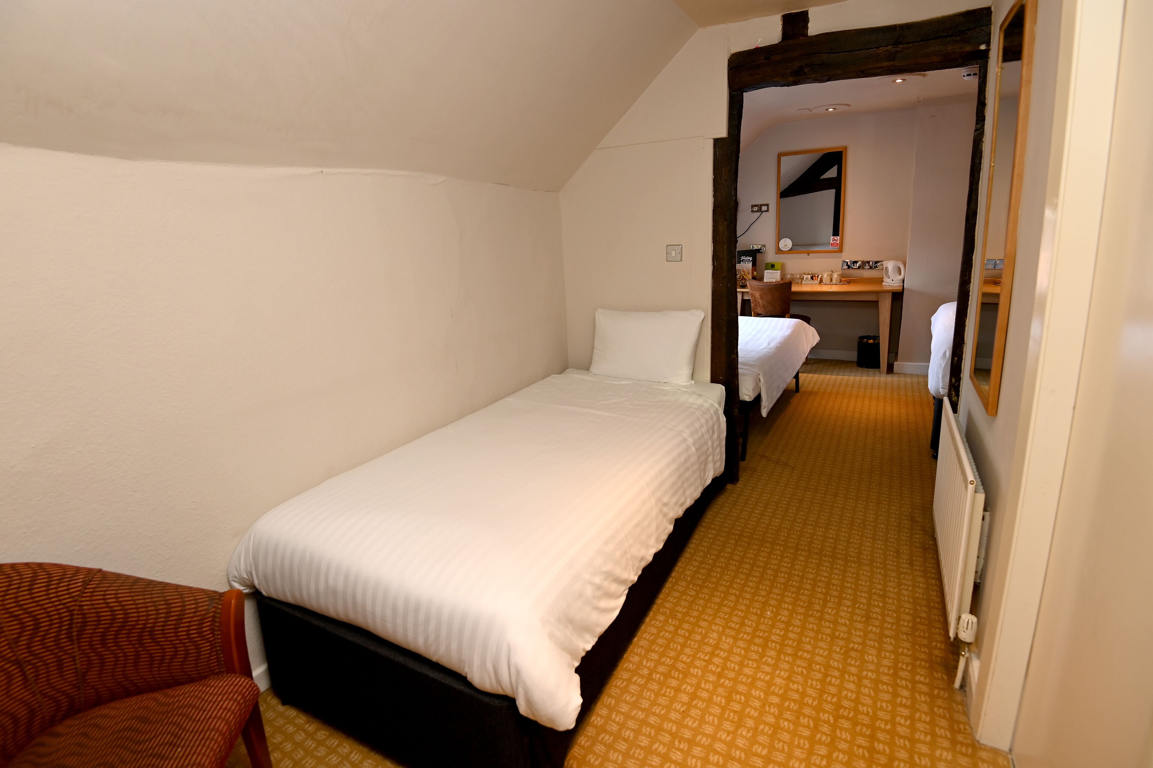 A small, single-bed room with a wooden floor, a wooden headboard, and a white bedspread. There is a wooden chair and a small table with a mirror and a kettle on it.