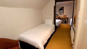 A small, single-bed room with a wooden floor, a wooden headboard, and a white bedspread. There is a wooden chair and a small table with a mirror and a kettle on it.