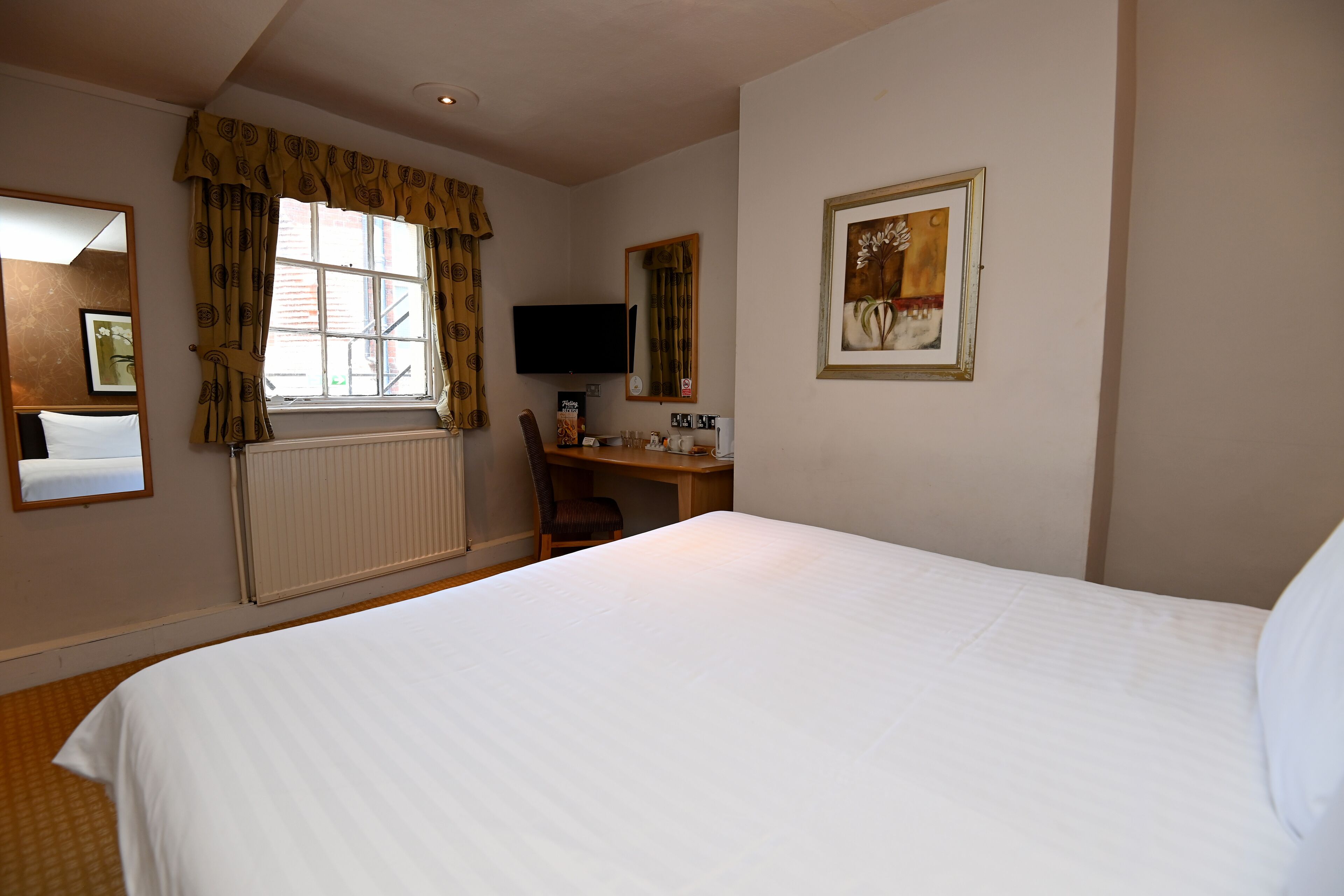 Een hotelkamer met een groot bed, een bureau met stoel, een televisie, een ingelijste foto aan de muur, en een raam met gordijnen.