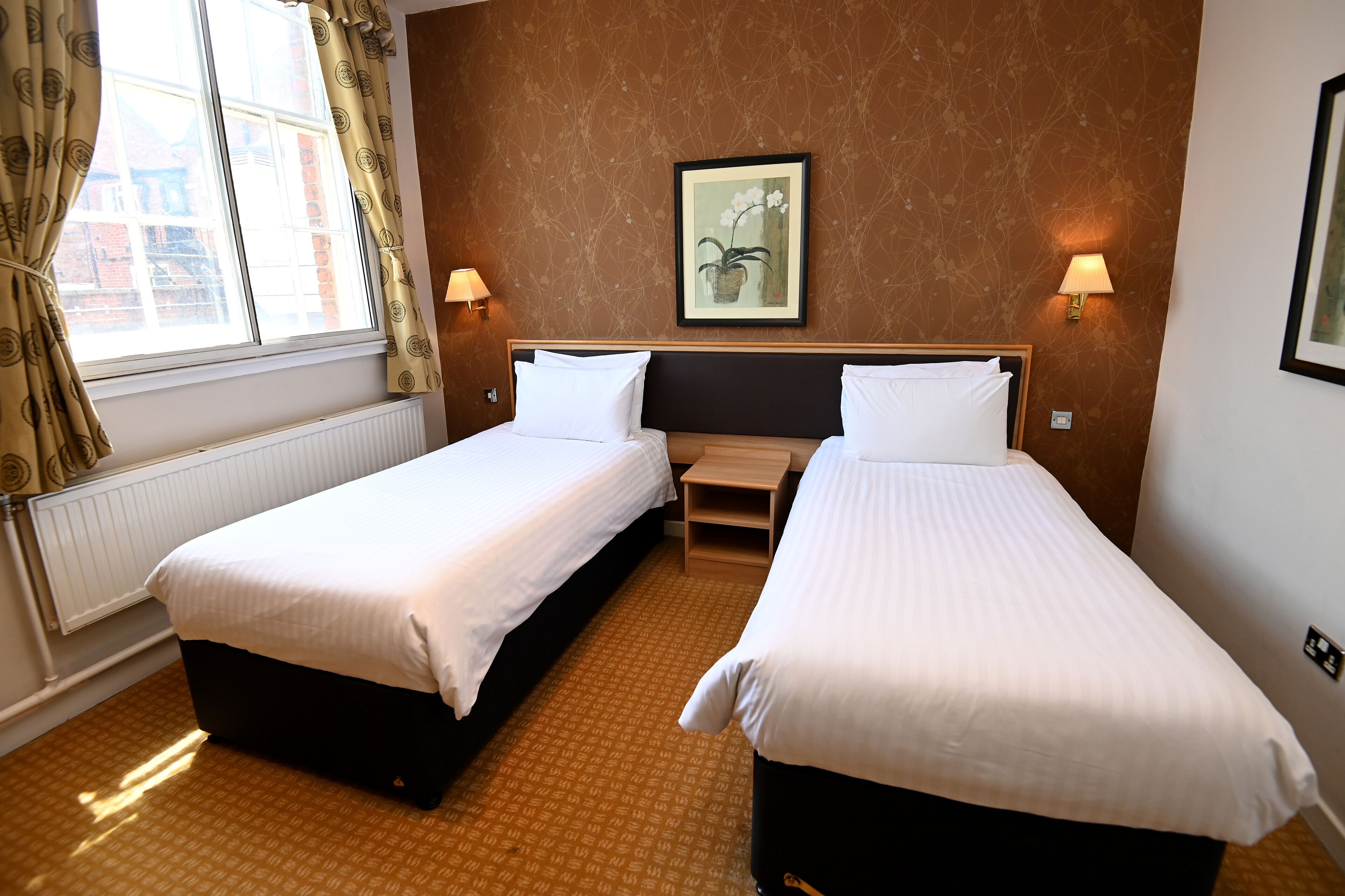 A hotel room with two single beds, a framed artwork on the wall, and a window with curtains.