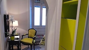 A hallway with a yellow chair, a lamp, and a desk with a plant. There is a window with blinds and a door leading to a room with a green wardrobe. - The George Of Stamford (Stamford)
