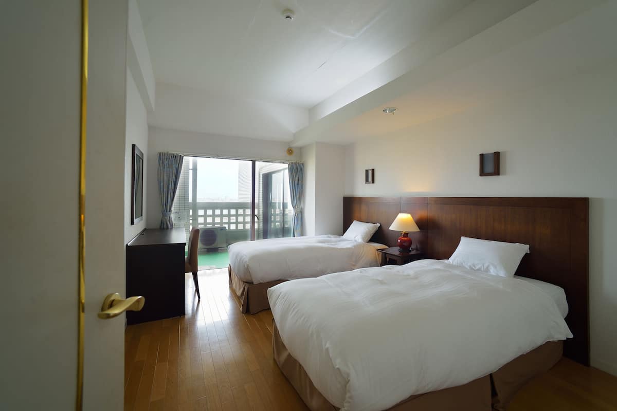 A hotel room with two beds, a desk, and a view of the outdoors.