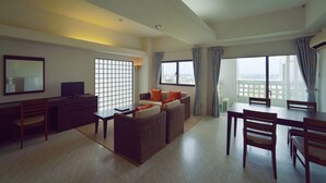 A spacious living area with a sofa set, a dining table, and chairs. There is a TV on a stand, a desk with a chair, and large windows with curtains. - Hotel Royal Marine Palace Ishigakijima (Ishigaki)
