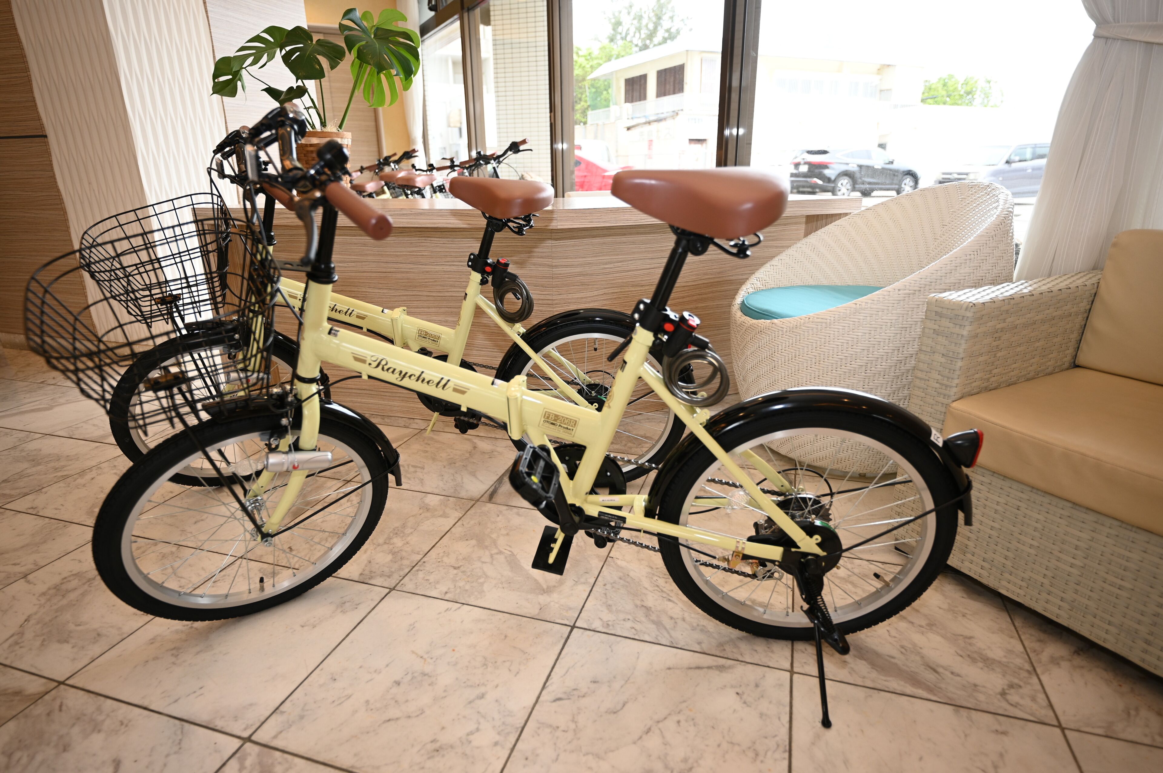 A yellow bicycle with a basket and a brown seat, positioned next to a wicker sofa in a room with tiled flooring and a large window.