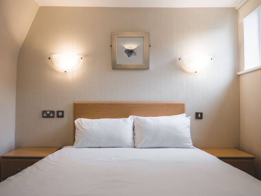 A neatly made bed with white linens, two bedside tables, and a wall-mounted light fixture above it.