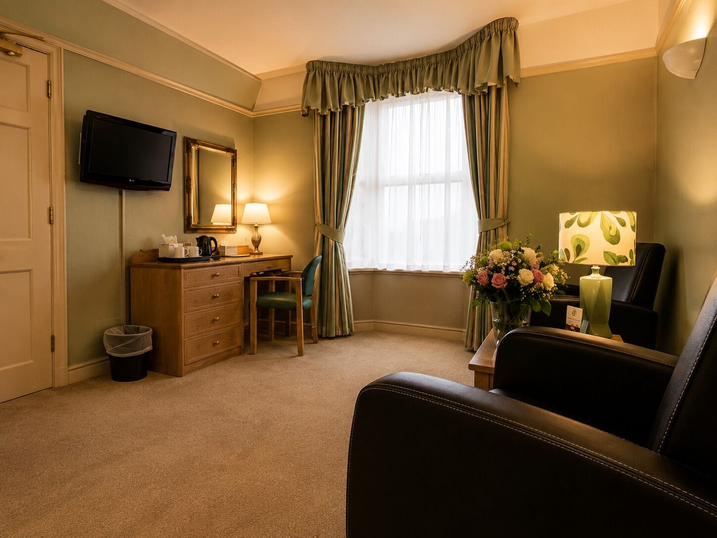 A hotel room with a flat-screen TV, a wooden desk with a lamp, a chair, a large window with curtains, and a vase of flowers.