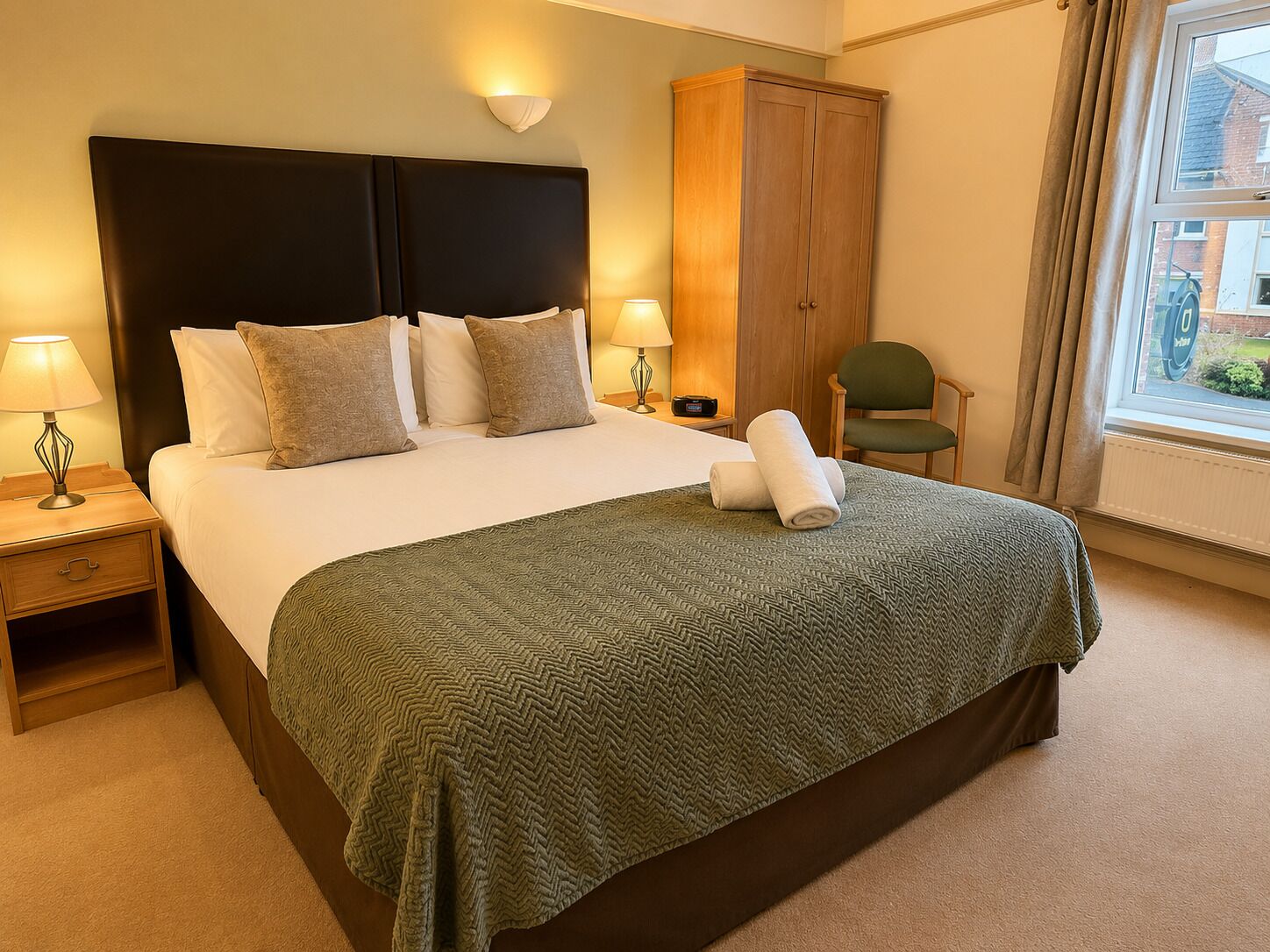 Habitación de hotel con una cama grande, dos lámparas de noche, una manta verde, un armario de madera y una silla.