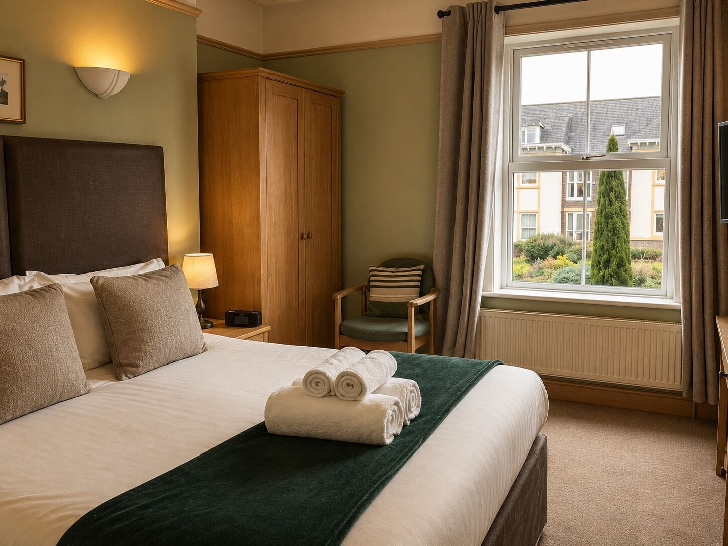 Ein Hotelzimmer mit Bett, Kleiderschrank, Stuhl, Lampe und Fenster mit Blick auf Gebäude und Grünflächen.