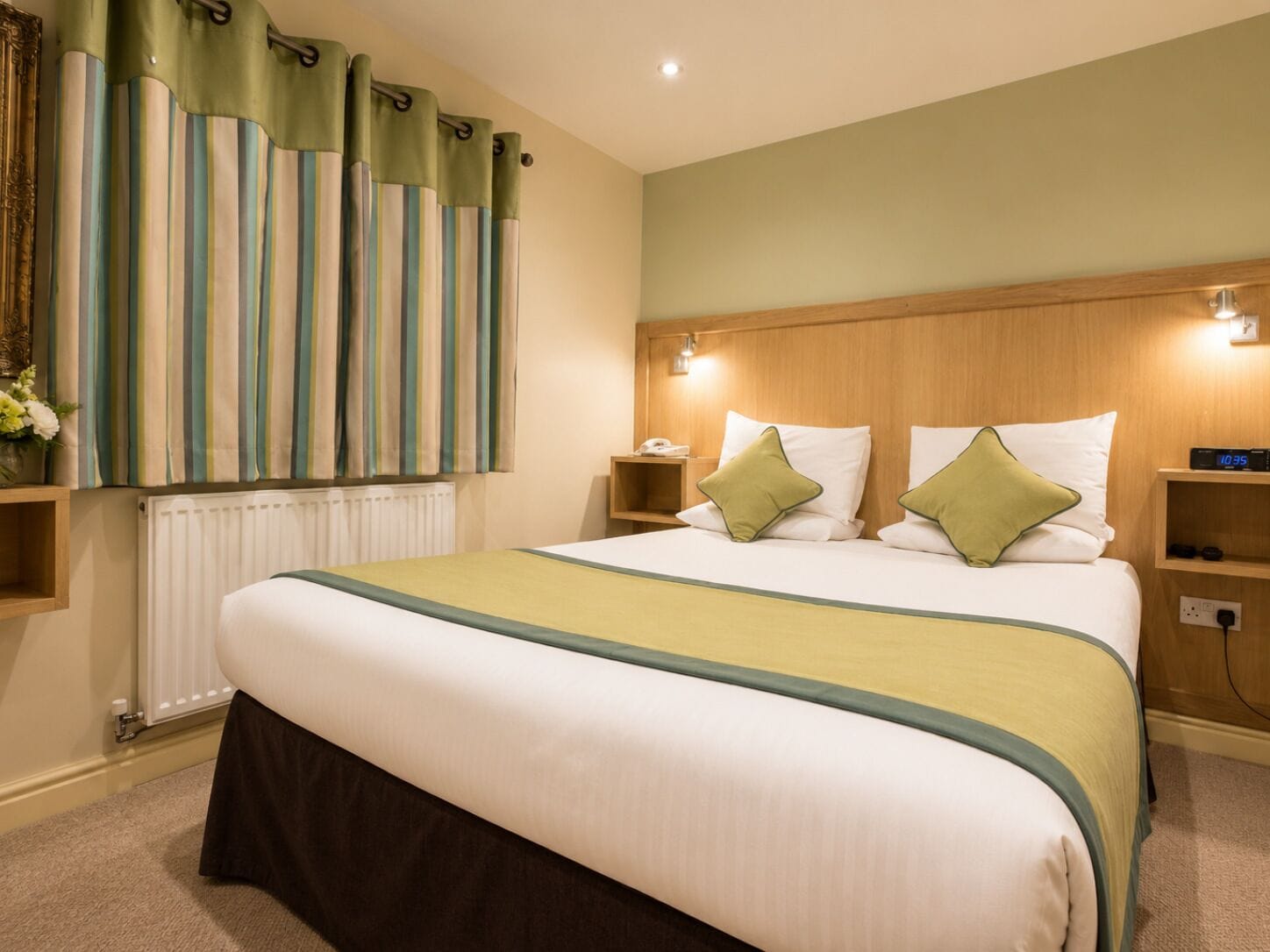 Een hotelkamer met een groot bed, twee nachtkastjes, een wandlamp en een raam met gordijnen.