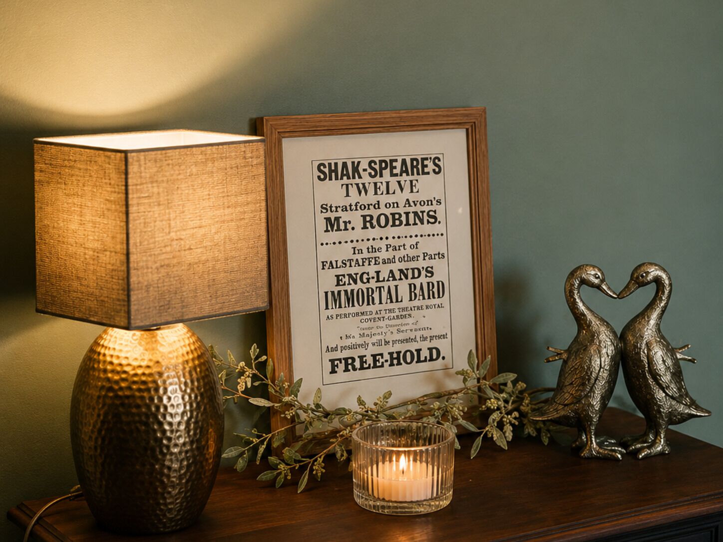 A framed sign with text, a lamp, a candle, and two duck figurines on a wooden surface.