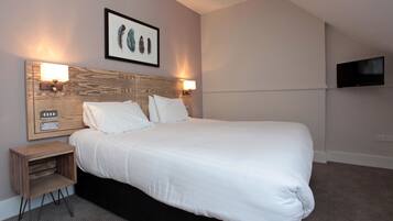 A neatly made bed with white linens, a headboard with a wooden panel design, two wall-mounted lamps, a small wooden nightstand with drawers, and a flat-screen TV mounted on the wall.
