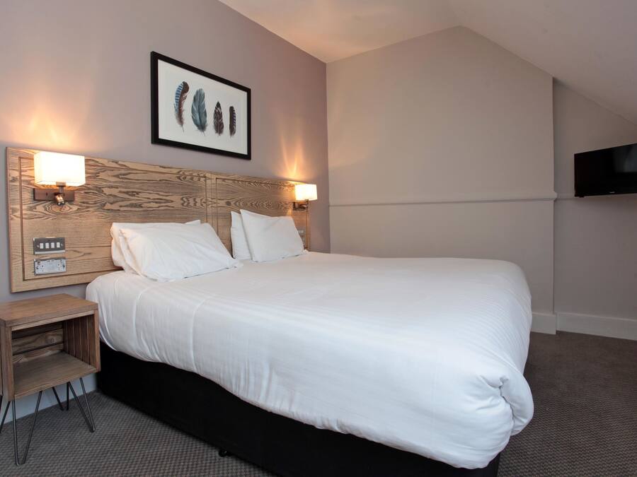 A neatly made bed with white linens, a headboard with a wooden panel design, two wall-mounted lamps, a small wooden nightstand with drawers, and a flat-screen TV mounted on the wall.