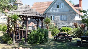 A stone building with a thatched roof structure, surrounded by greenery and picnic tables. - Bear Inn, Somerset by Marston's Inns (Street)