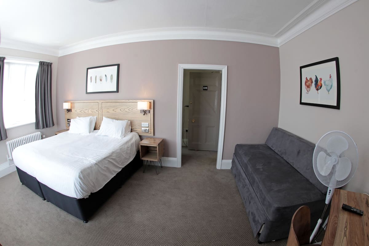 Una habitación de hotel con una cama grande, un sofá, un ventilador de pie y una puerta que da a otra habitación.