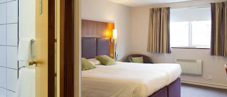 A hotel room with a bed, a bedside table, a lamp, a window with curtains, and a wall-mounted heater.