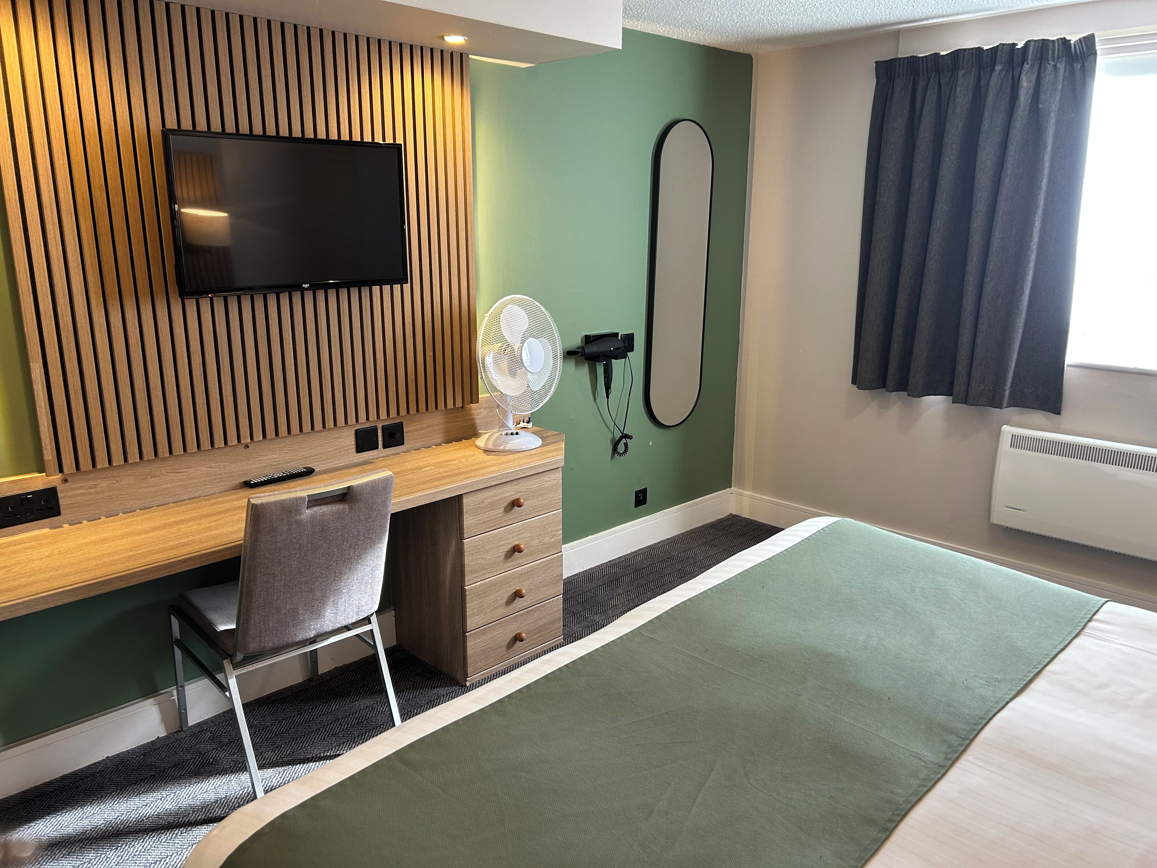 A hotel room with a wooden desk, a flat-screen TV, a standing fan, a mirror, a bed with a green bedspread, and a window with curtains.