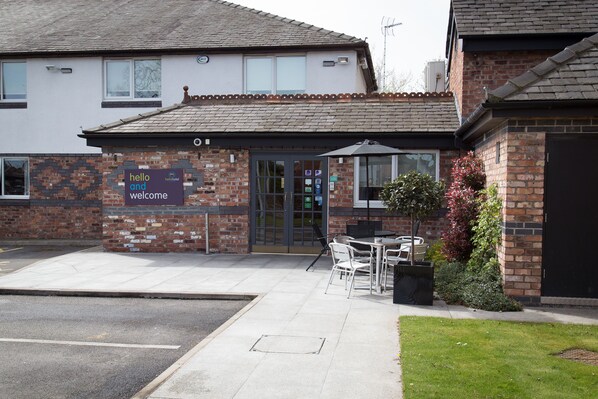 A hotel entrance with a 'hello and welcome' sign, outdoor seating, and a brick facade.