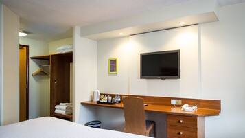 Une chambre d’hôtel comprenant un lit, un bureau avec une chaise, une télévision et une armoire avec des vêtements suspendus.
