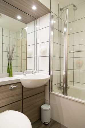 Un baño moderno con un espejo grande, un lavamanos blanco, una mampara de cristal para la ducha y un pequeño cubo de basura.
